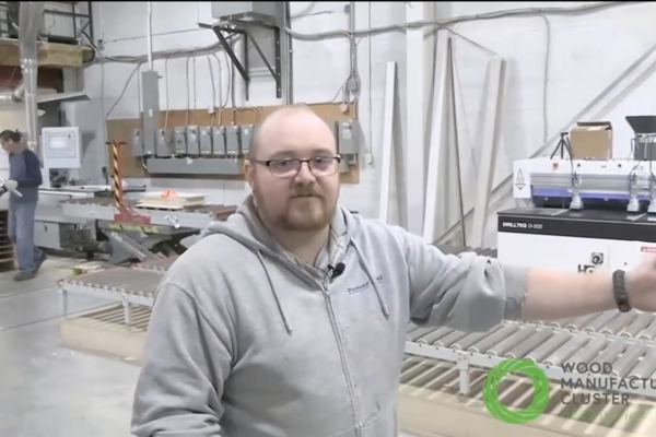 Rob Clare standing inside Diamond CNC shop with various pieces of equipment and a worker in background. He has his arm raised as he gives his tour of the shop.