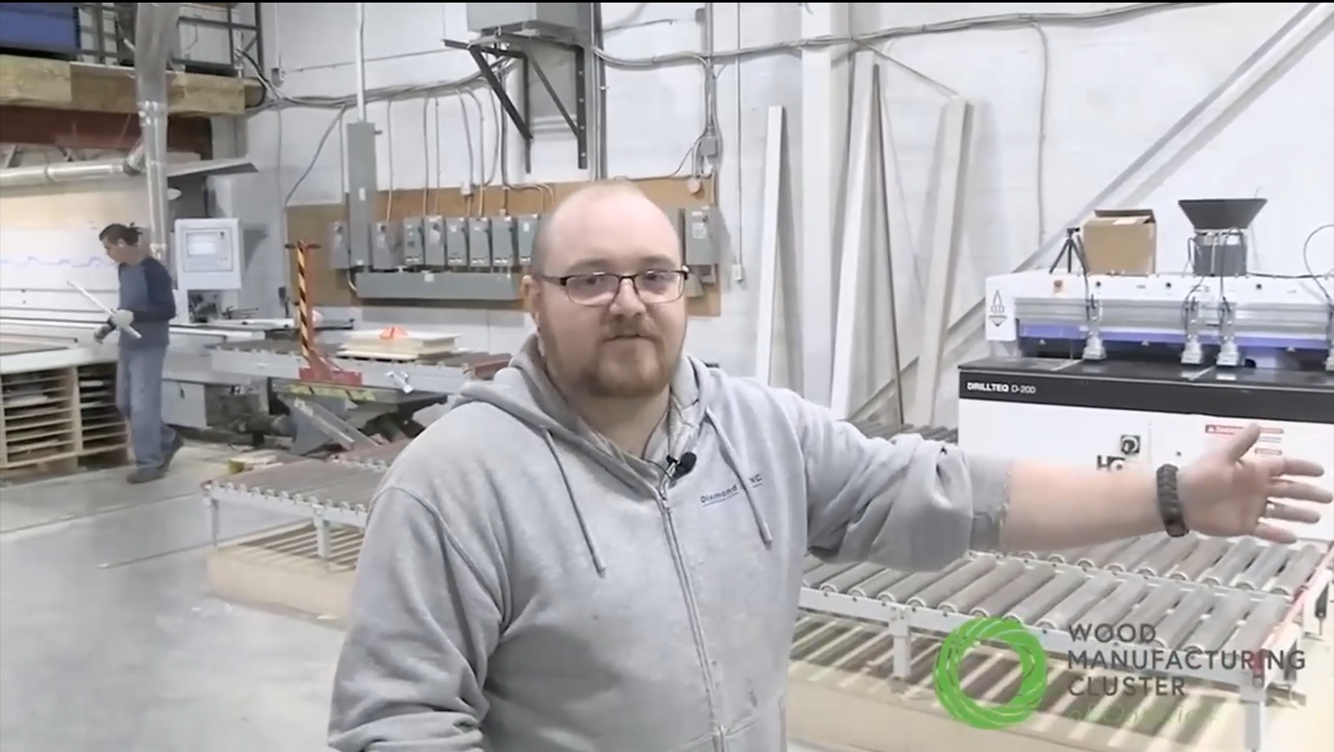 Rob Clare standing inside Diamond CNC shop with various pieces of equipment and a worker in background. He has his arm raised as he gives his tour of the shop.